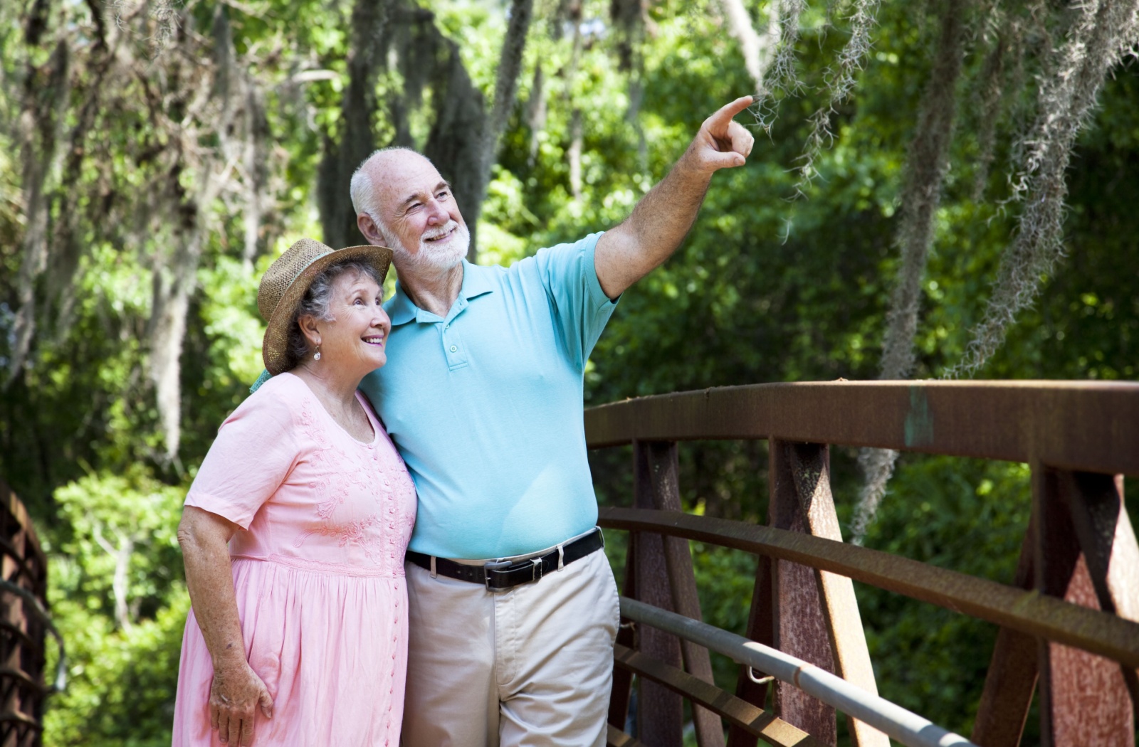 An older adult couple standing on a bridge in a forest and smiling as the man points at something.