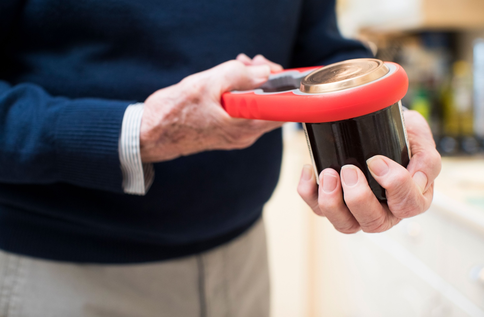 A close-up of an older adult's hands holding a jar in his left hand and opening it with the help of a jar opener in his right hand.