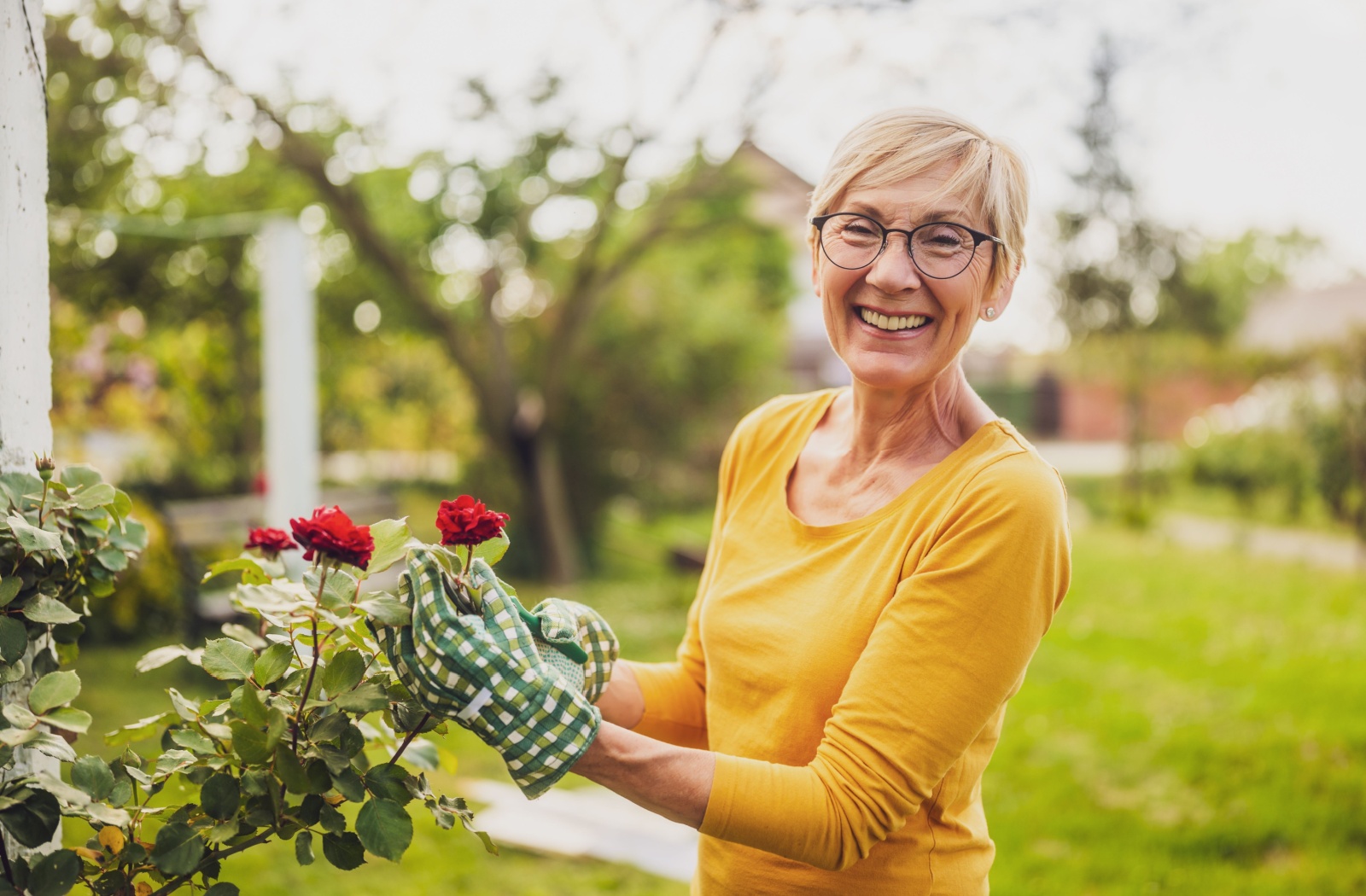 An older adult woman with short hair working on her rose garden with a big smile on her face