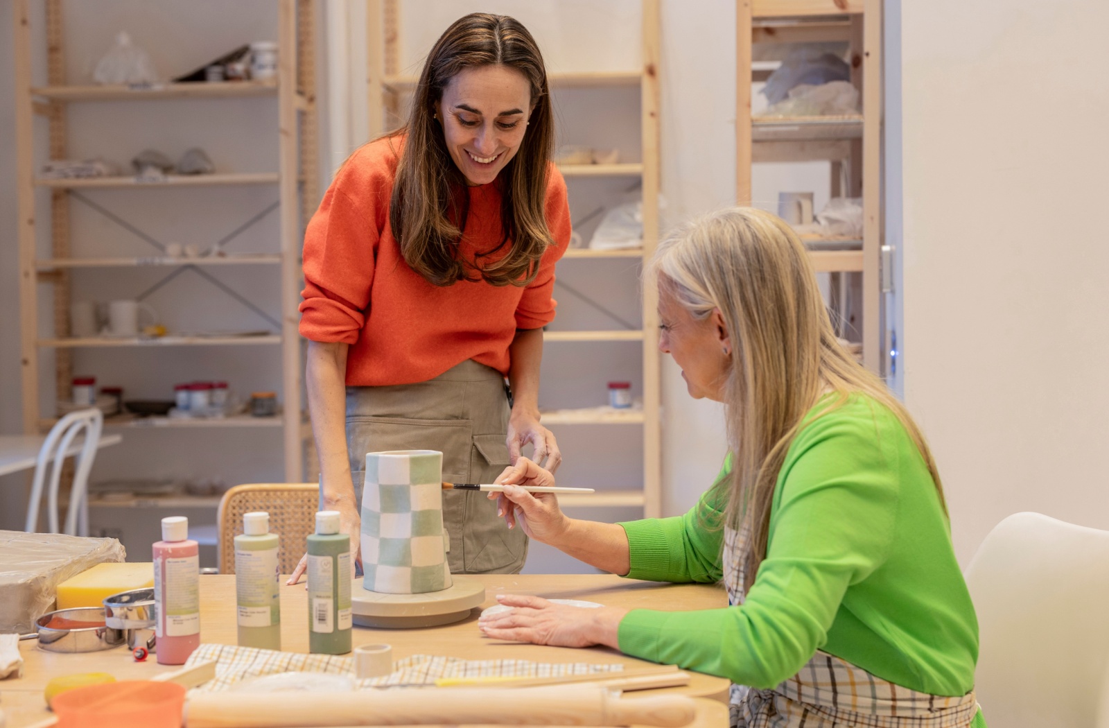 Two women working in a pottery studio. One looks on and smiles while the other paints a blue and white checkered vase.