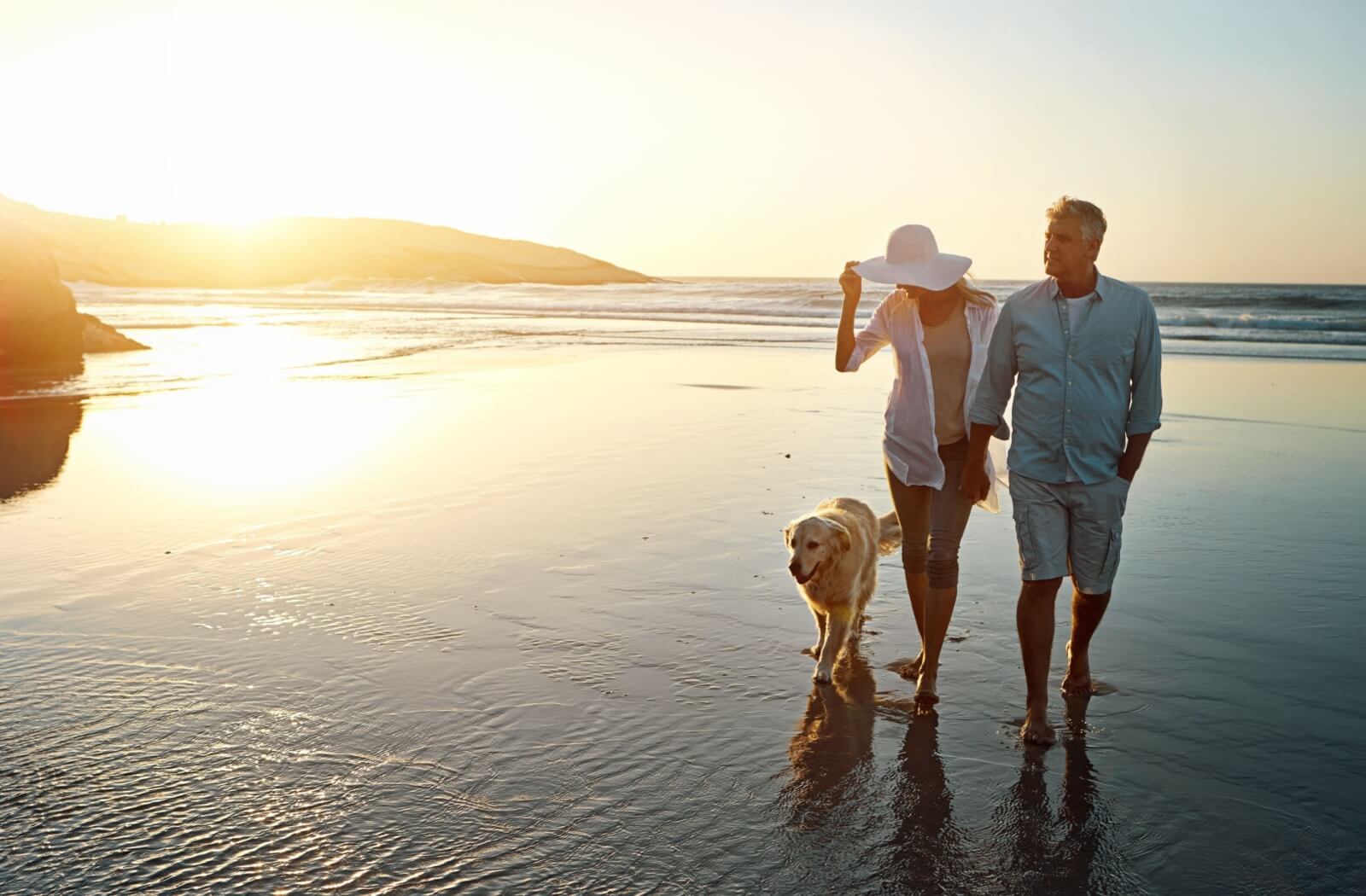 A retired couple walking along the beach with their dog at sunset.